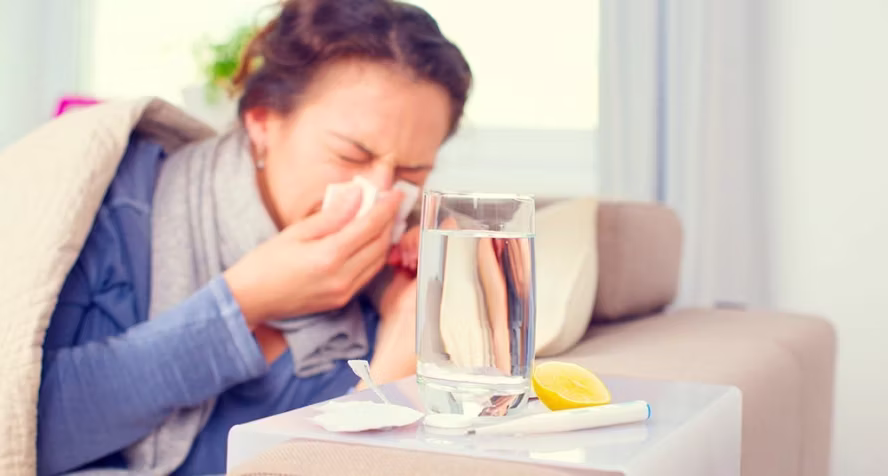 A woman wrapped in a blanket sits on a sofa blowing her nose into a tissue. A glass of water, a lemon slice, and medication are on a small table in the foreground